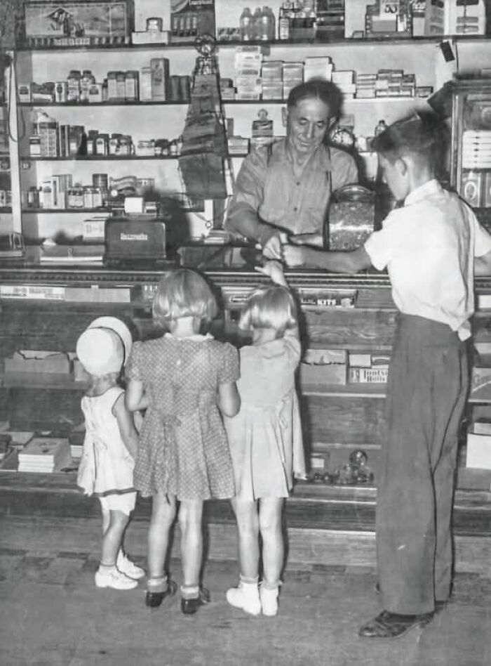 Niños comprando en una tienda antigua, atendidos por un tendero detrás del mostrador, evocando días pasados.
