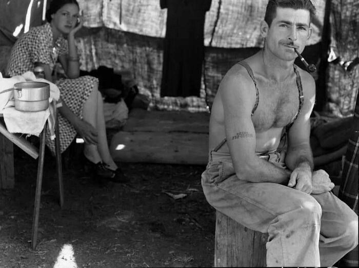 Hombre fumando pipa sentado en un taburete, junto a una mujer en un campamento, evocando días pasados históricos.