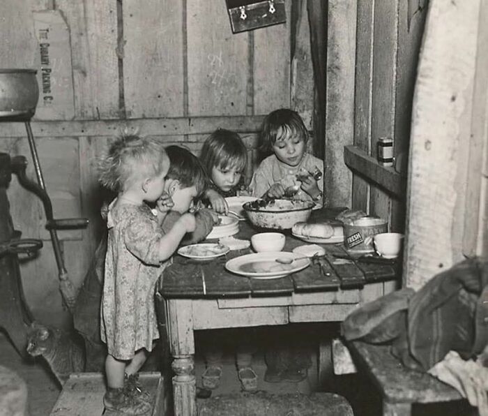 Niños en mesa compartiendo comida en entorno rústico, ilustrando fotos históricas de días pasados.