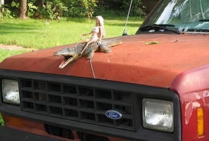 Muñeca sobre cocodrilo de juguete en capó de camioneta roja, ejemplo de imágenes raras y absurdas.