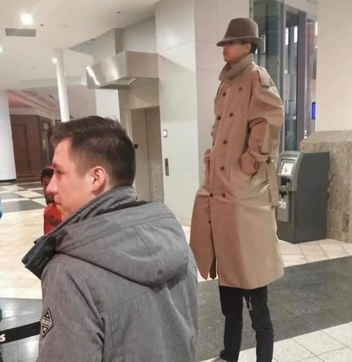 Hombre con abrigo largo parado sobre los hombros de otro, creando una imagen rara y absurda en un centro comercial.