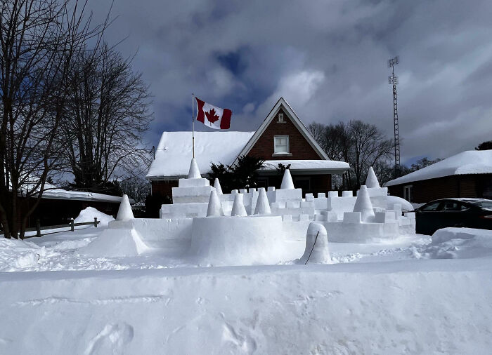 Casa nevada en Canadá con bandera canadiense ondeando, rodeada de esculturas de nieve.
