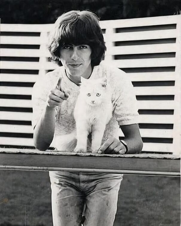 Desconocida foto histórica de un hombre joven con un gato blanco, señalando hacia la cámara frente a una cerca.