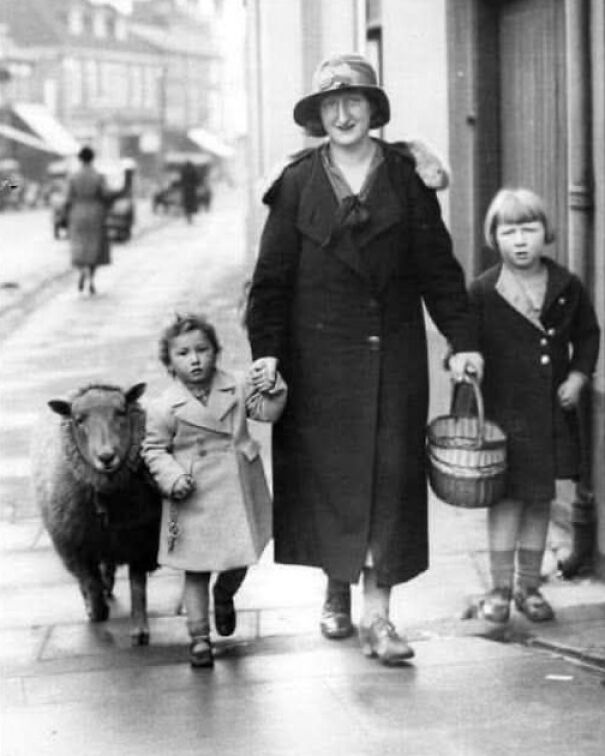 Mujer paseando con dos niños y una oveja en una calle antigua, evocando recuerdos de días pasados.