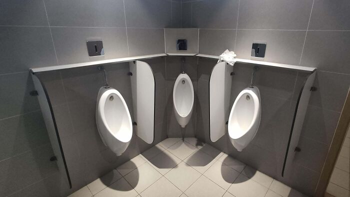 Urinales en esquina mal diseñados en un baño público, mostrando problemas de privacidad. Diseño curioso y poco funcional.