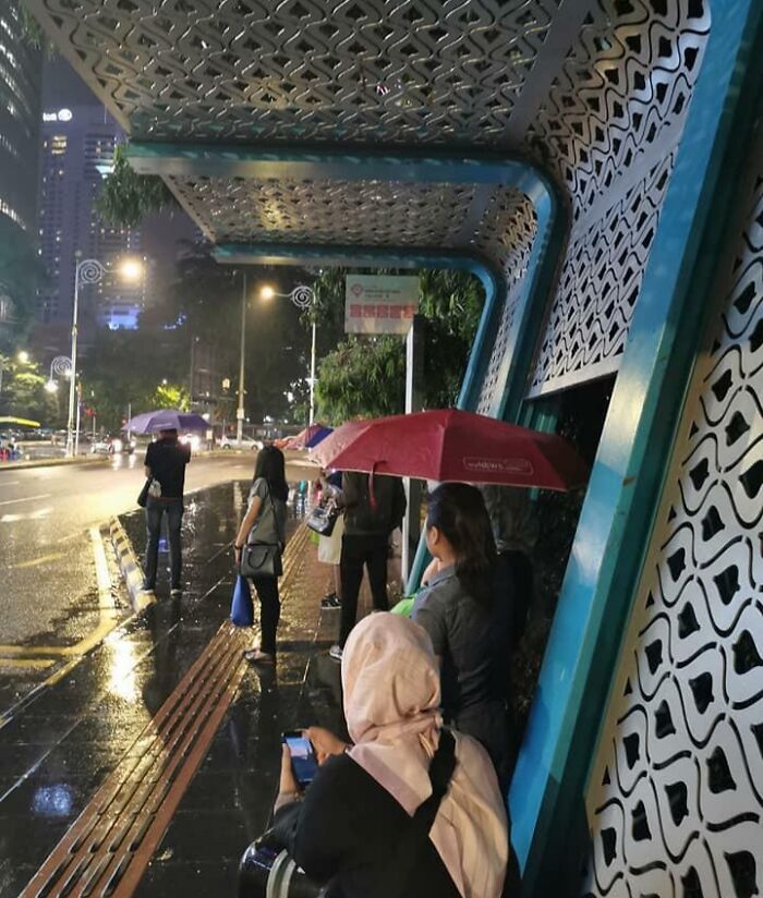 Personas con paraguas bajo una estructura de diseño complejo en una noche lluviosa, esperando en la acera.