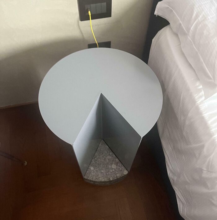 Mesa de diseño inusual junto a una cama, mostrando un compartimiento triangular y cable amarillo conectado al enchufe.