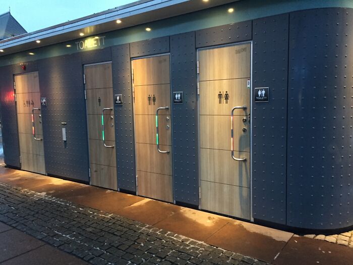 Baños públicos innovadores con diseño moderno en una calle adoquinada, mostrando detalles ingeniosos en el exterior.