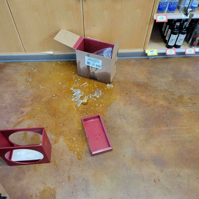 Caja rota con botellas quebradas y líquidos derramados en el suelo de una tienda, mostrando un mal día en el trabajo.