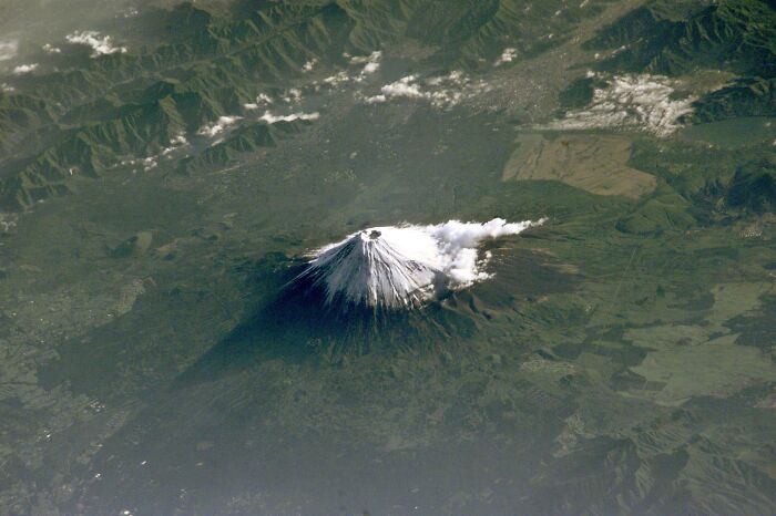 Perspectiva aérea del Monte Fuji cubierto de nieve, rodeado de vegetación y montañas.