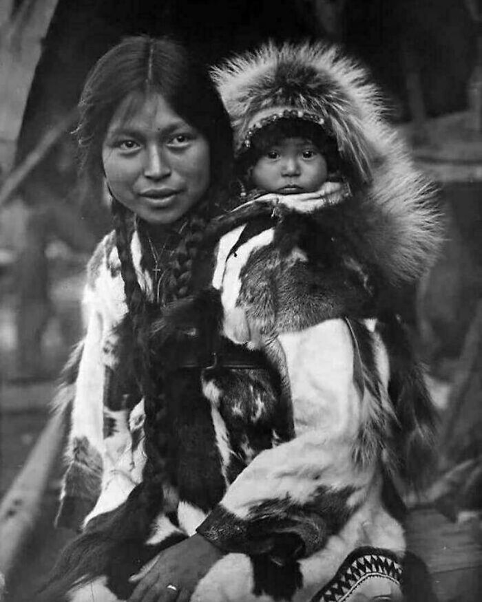 Mujer indígena con niño en espalda vistiendo ropa tradicional de piel en foto histórica fascinante en blanco y negro.