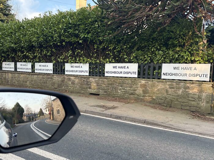 Carteles pasivo-agresivos en una valla anuncian disputa vecinal, vistos desde un coche en movimiento.
