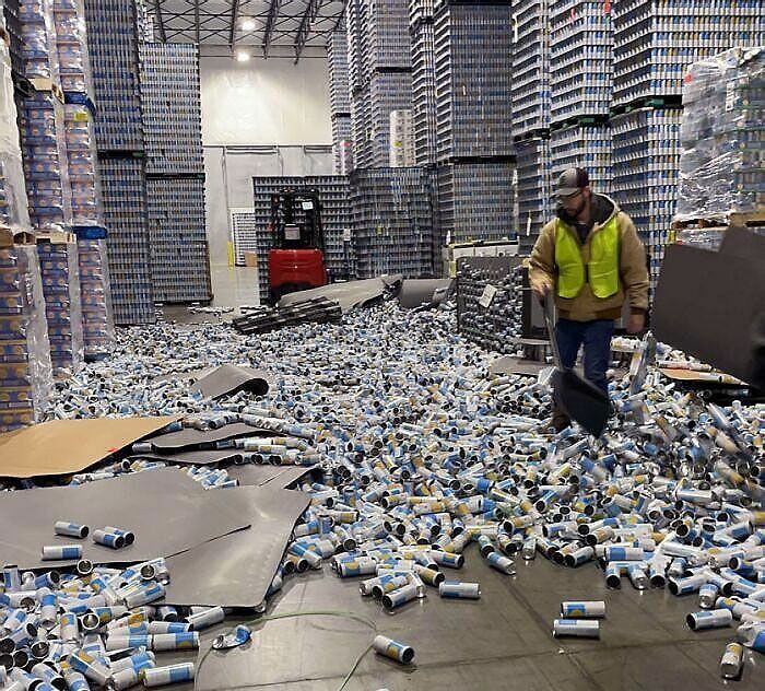 Trabajador con chaleco amarillo limpiando latas caídas en un almacén desordenado, día difícil en el trabajo.