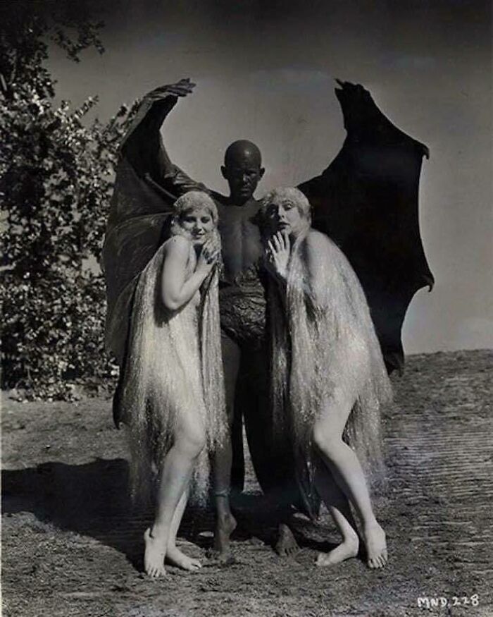 Imagen inquietante del pasado: hombre disfrazado de criatura con alas oscuras junto a dos mujeres con vestidos brillantes.