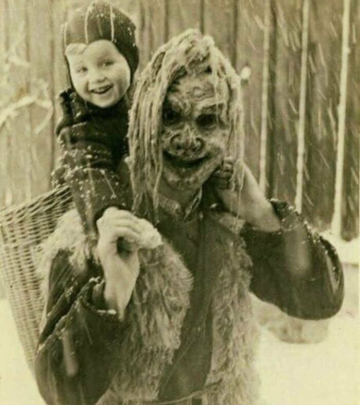 Persona disfrazada con máscara inquietante cargando a un niño sonriente en la nieve, una escena del pasado.