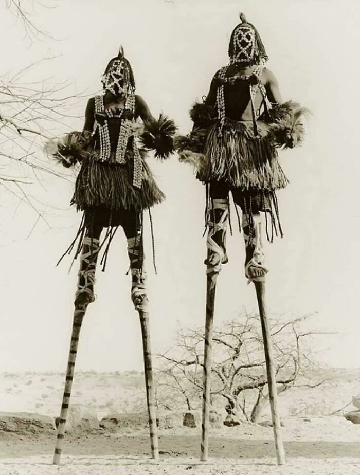 Dos personas en zancos con disfraces tribales, representando imágenes inquietantes del pasado.
