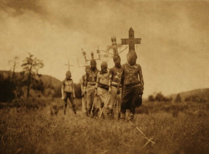 Personas con máscaras y cruces, representando imágenes inquietantes del pasado en un campo abierto.
