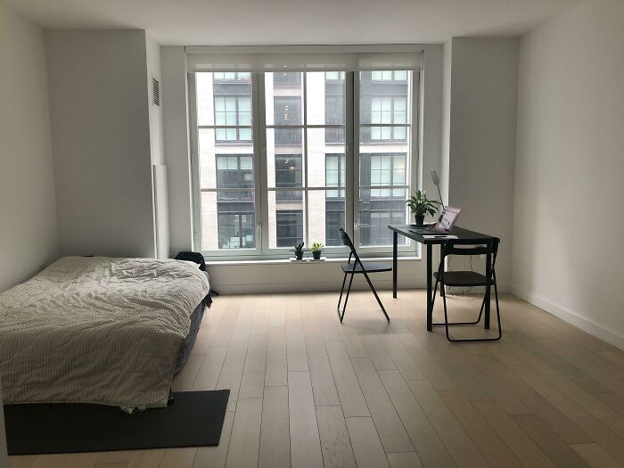 Habitación minimalista con cama sencilla y escritorio negro frente a ventana grande, reflejando tendencias de interiorismo.