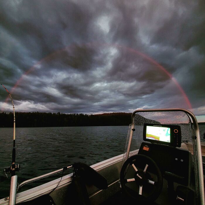 Barco bajo un arco iris en el lago con cielo nublado, creando una escena surrealista.