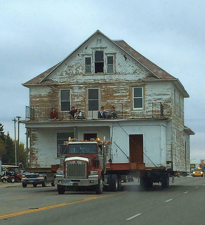 Casa antigua siendo transportada en un camión grande por una carretera. Personas sentadas en el balcón observan.
