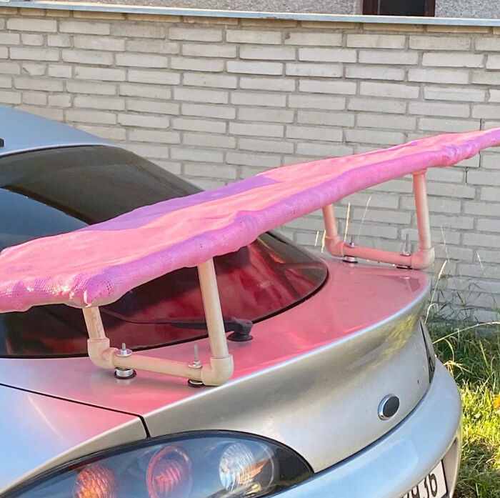 Alerón casero en auto plateado hecho con malla rosa y tuberías, reflejando intento fallido de reparación.