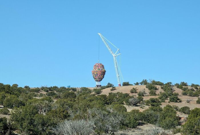 Grúa levantando una gran escultura en forma de cabeza hecha de globos, rodeada de paisaje natural.