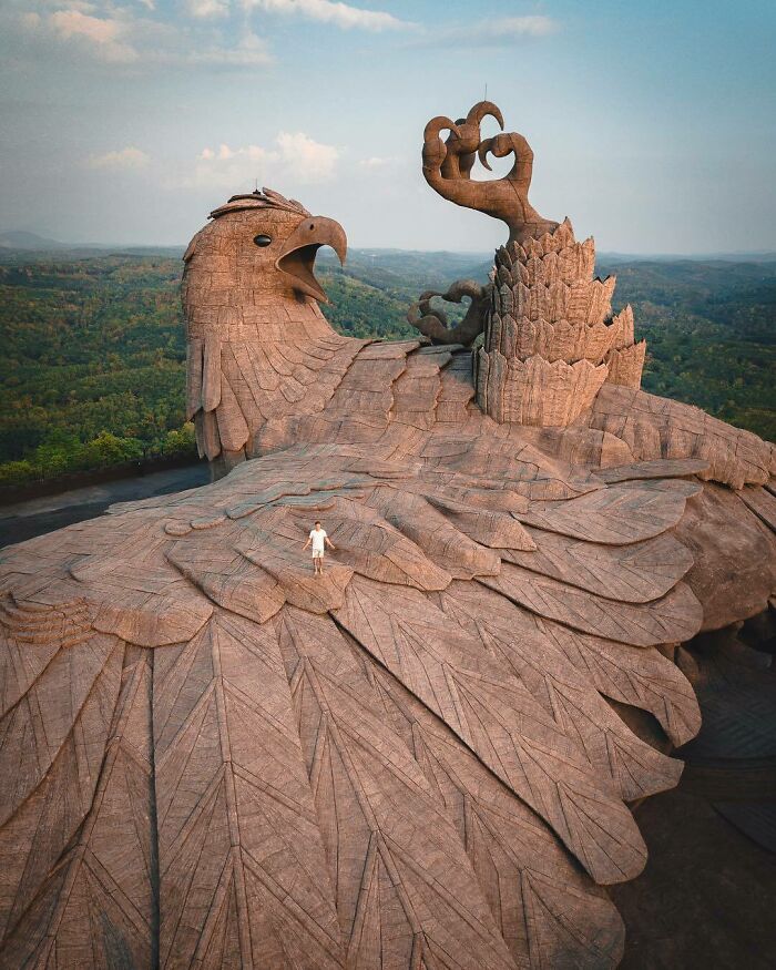 Un hombre pequeño sobre una gigantesca escultura de águila de piedra, destacando lo surrealista de la escena.