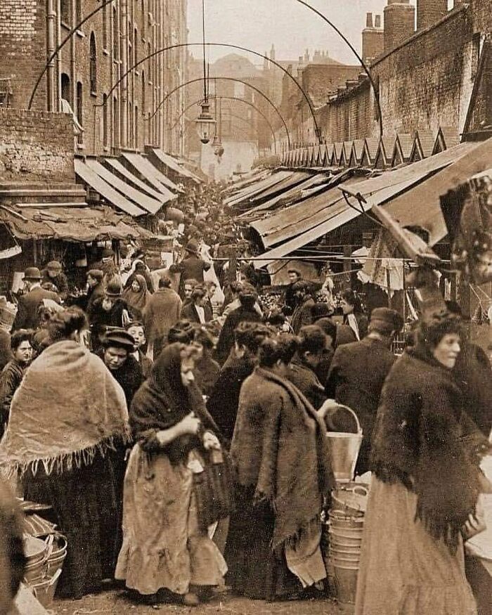 Multitud en un mercado histórico al aire libre, con sombrillas y edificios antiguos en un entorno urbano del pasado.