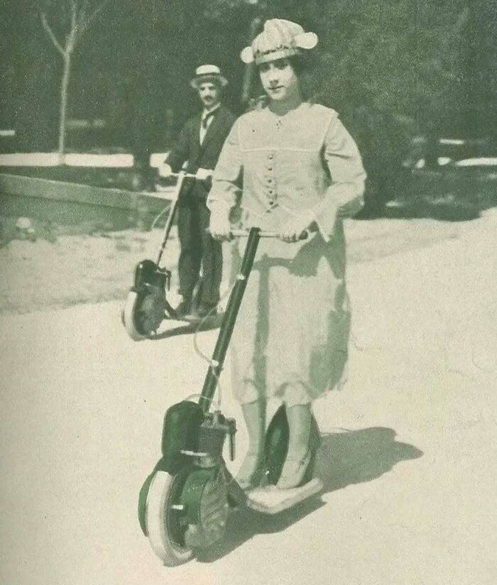 Mujer en patinete antiguo con vestido y sombrero, seguida por hombre. Fotografía histórica.