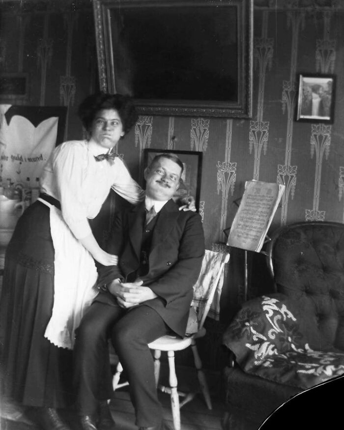 Pareja en foto histórica, ella de pie con blusa clara y él sentado con traje oscuro, en un salón antiguo.
