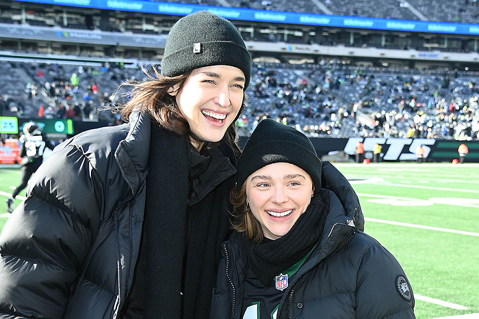 Pareja de celebridades ultra privada en un estadio de fútbol americano, sonriendo y vistiendo ropa de invierno.