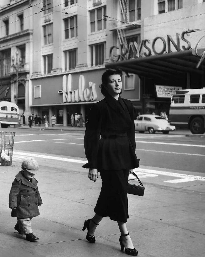 Mujer elegante caminando con un niño en la calle de una ciudad histórica, reflejando moda del pasado.