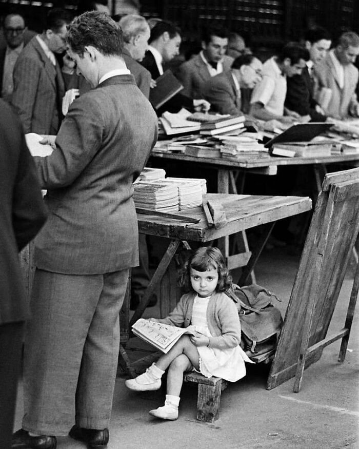 Niña con un cómic en un mercado de libros antiguo, rodeada de adultos absortos en lectura, escena histórica.