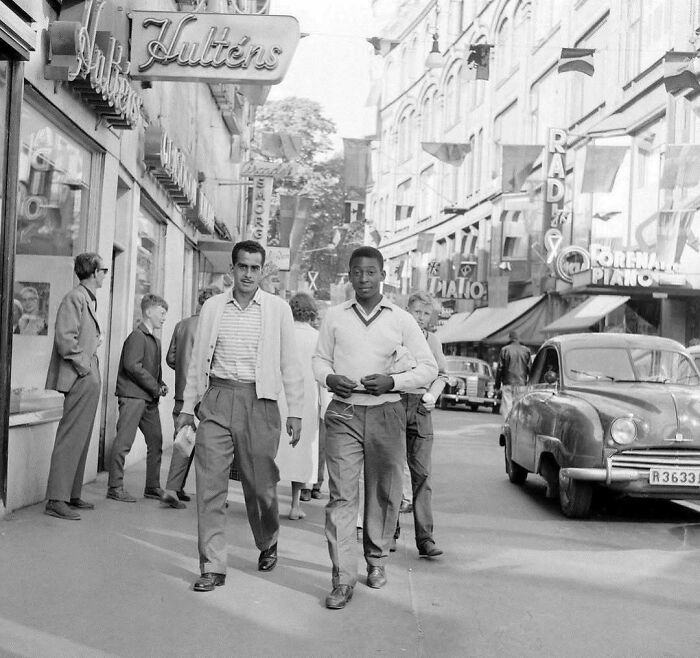 Dos hombres caminan por una concurrida calle histórica, rodeados de letreros y coches antiguos.