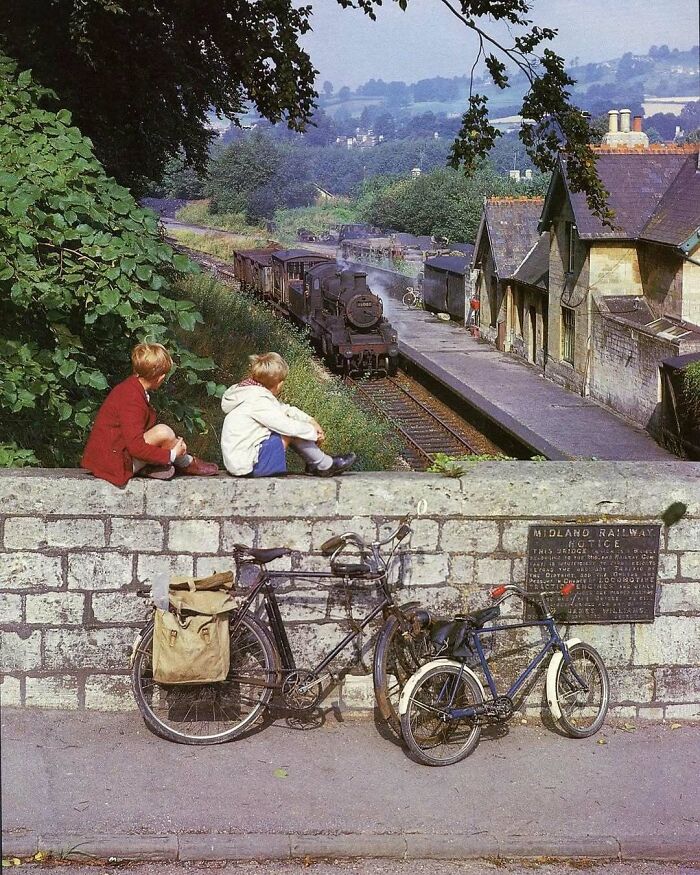 Dos niños observan un tren antiguo desde un muro, con bicicletas apoyadas, evocando fotos históricas.