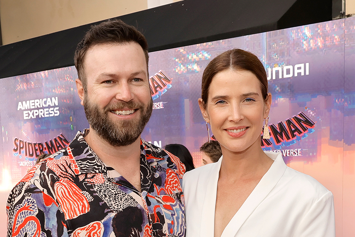 Pareja de celebridades en evento de estreno, luciendo looks elegantes frente a un cartel de Spider-Man.