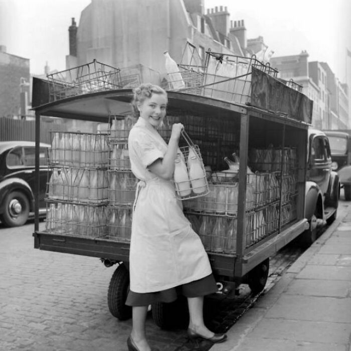 Mujer sonriente en uniforme junto a un camión de reparto de leche antiguo en una calle urbana.