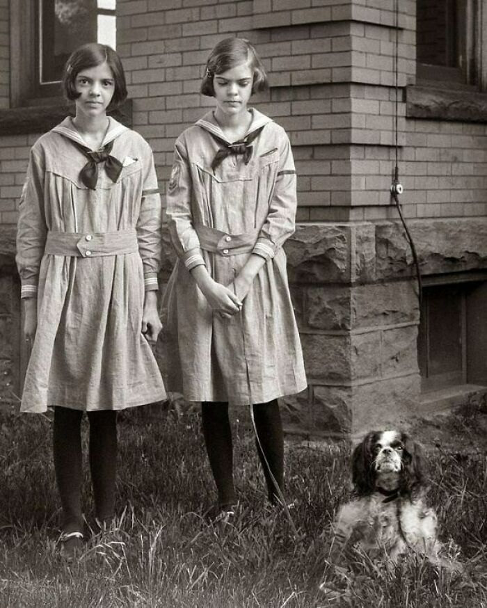 Dos niñas con vestido antiguo y un perro en un jardín, reflejando cambios sociales con el tiempo.