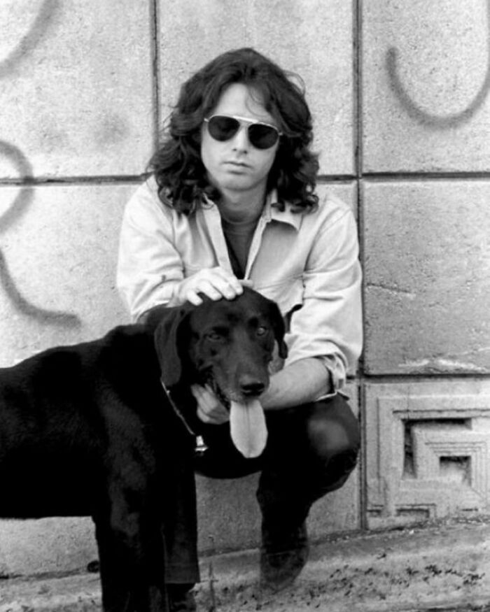 Persona con gafas de sol acariciando a un perro frente a una pared grafiteada; escena de foto antigua.
