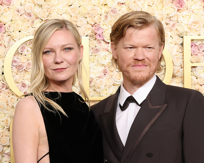 Pareja de celebridades privadas en evento, con vestido negro y traje oscuro frente a fondo de flores.