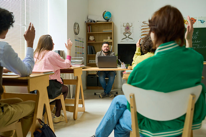 Estudiantes en clase levantan la mano mientras el profesor escucha, ilustrando experiencias infantiles en el aula.