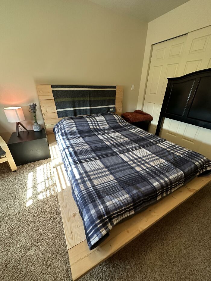Cama de madera minimalista con manta de cuadros en tendencia de interiorismo poco práctica y mesa de noche con lámpara.