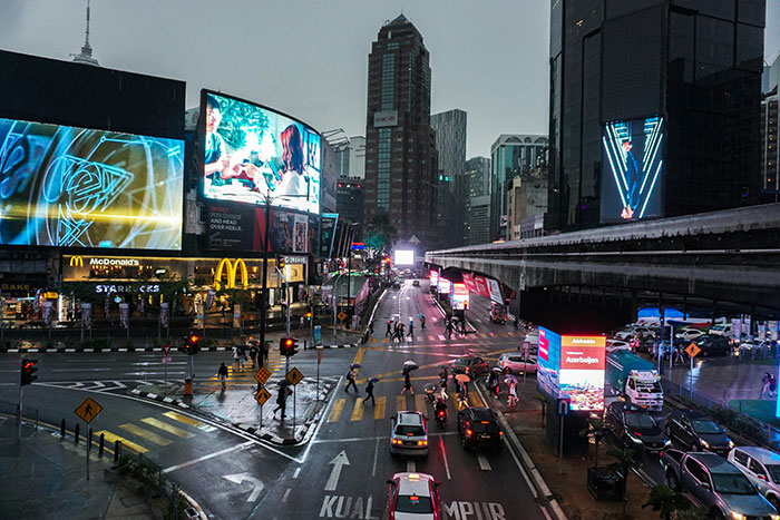 Cruce urbano concurrido en Kuala Lumpur con tráfico, peatones y carteles luminosos, reflejando un choque cultural.