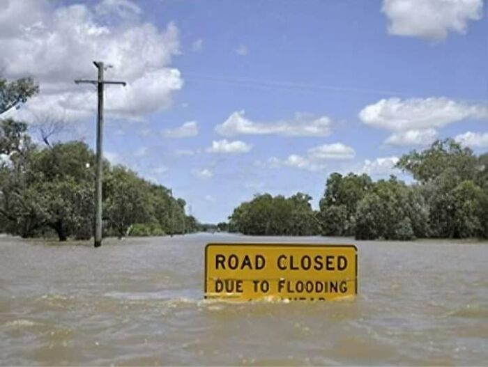Señal de aviso legal de carretera cerrada por inundación en zona rural con árboles y cielo nublado.