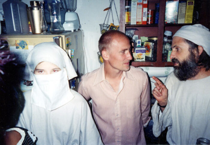 Personas conversando en una cocina abarrotada, incluyendo a una figura con vestimenta inusual. Foto inquietante.