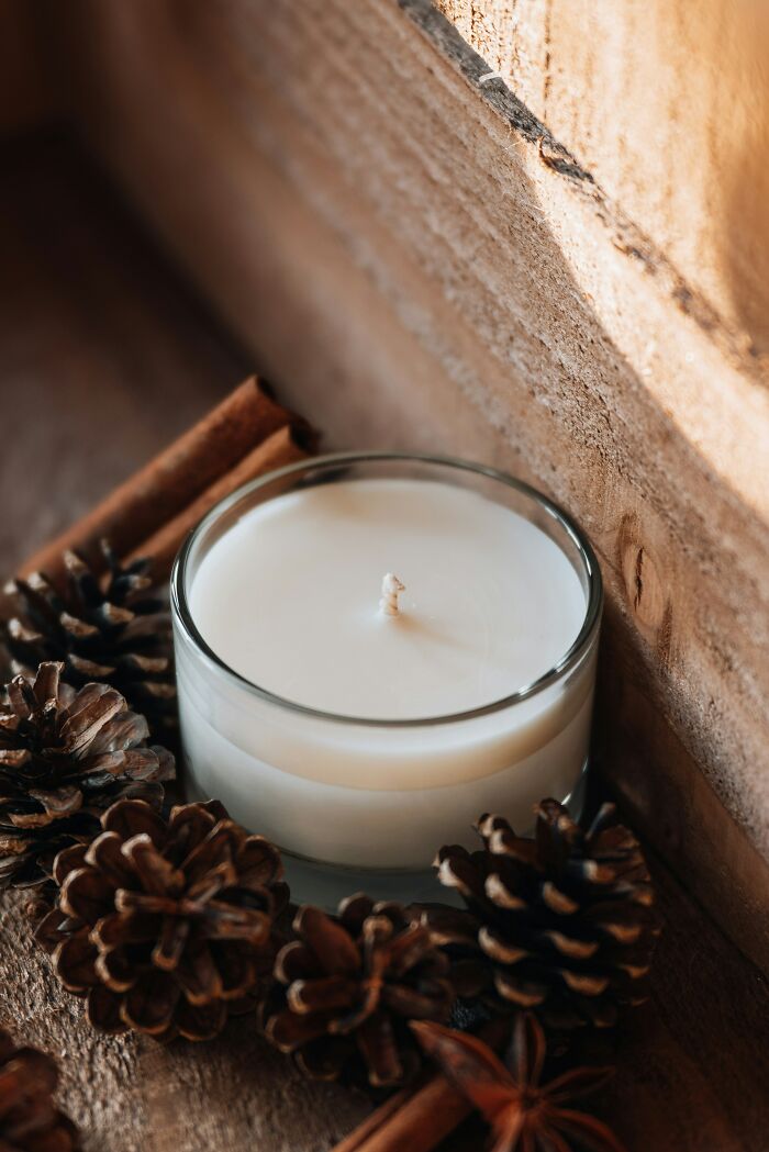 Vela blanca en un vaso de cristal sobre madera, rodeada de conos de pino y canela.