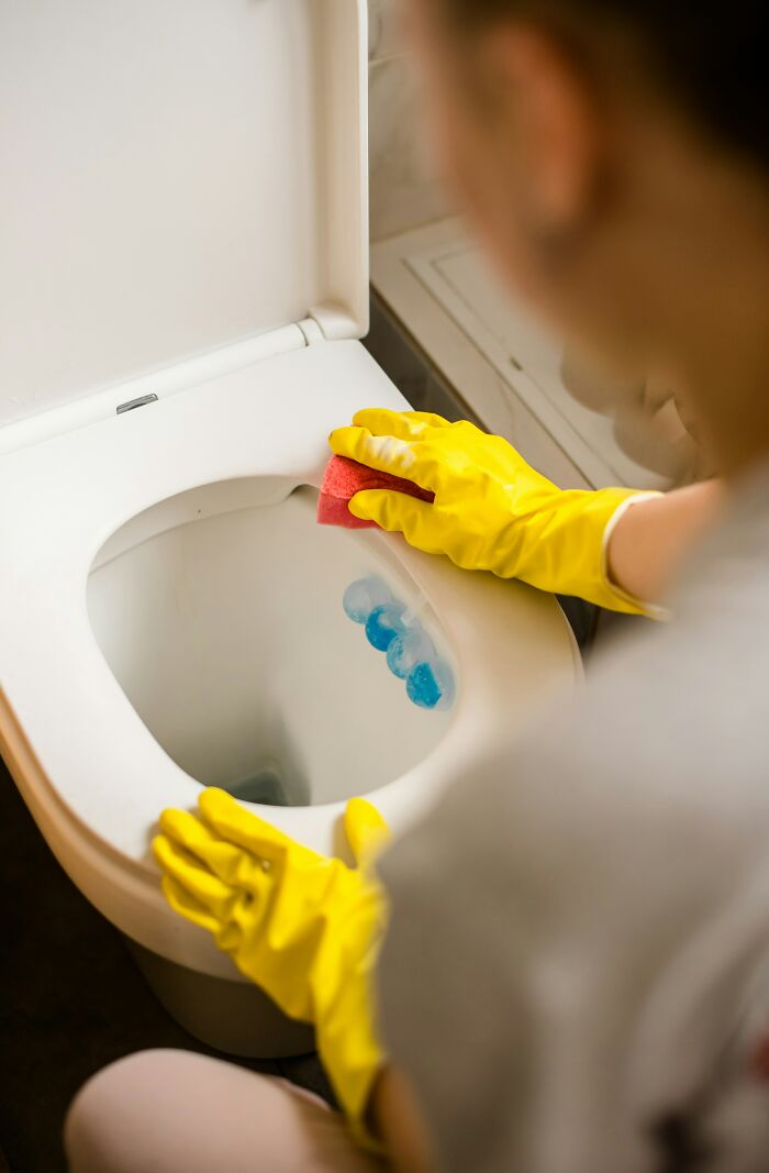 Persona usando guantes amarillos limpiando un inodoro con una esponja roja.
