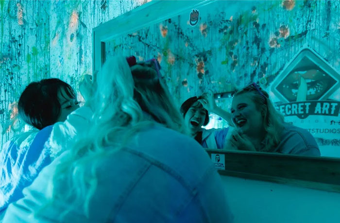 Mujeres riendo frente al espejo en una habitación iluminada de azul.