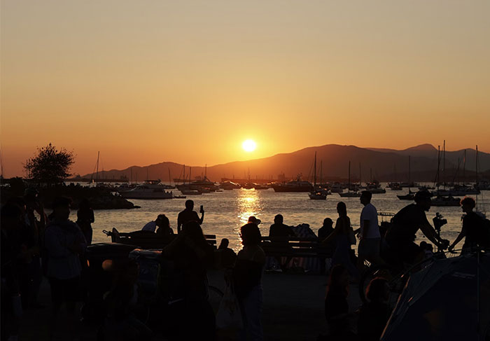 Personas en un puerto al atardecer, siluetas coinciden con el fondo del paisaje, creando una escena sorprendente.