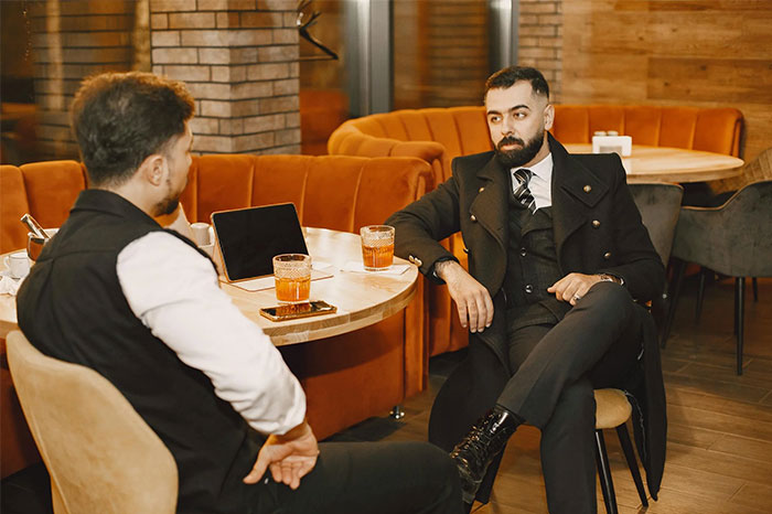 Dos hombres discutiendo en un café, con tazas y un portátil sobre la mesa, creando una coincidencia visual asombrosa.